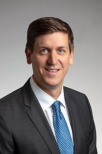 A person wearing a suit jacket, dress shirt, and tie, facing the camera against a plain background.