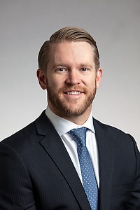 A person wearing a suit jacket, dress shirt, and tie, facing the camera against a plain background.
